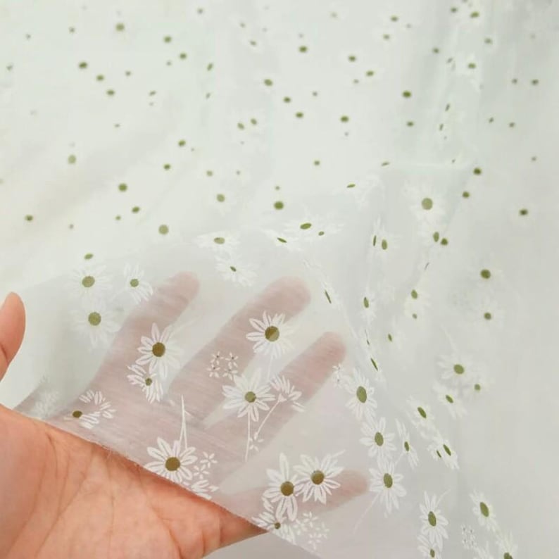 May include: A sheer white fabric featuring a repeating pattern of small white daisy flowers with olive green centers. The lightweight, translucent material is draped to show the delicate floral print and the fine texture of the textile.