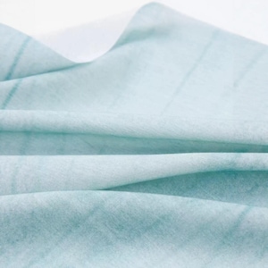 K&ouml;nnte beinhalten: Nahaufnahme eines weichen, hellblauen Stoffes mit dezenten, dunkleren blauen Linien. Der Stoff ist drapiert und gefaltet, wodurch ein strukturiertes Aussehen entsteht. Das Material wirkt leicht und eignet sich m&ouml;glicherweise f&uuml;r Kleidung oder Heimdekoration.
