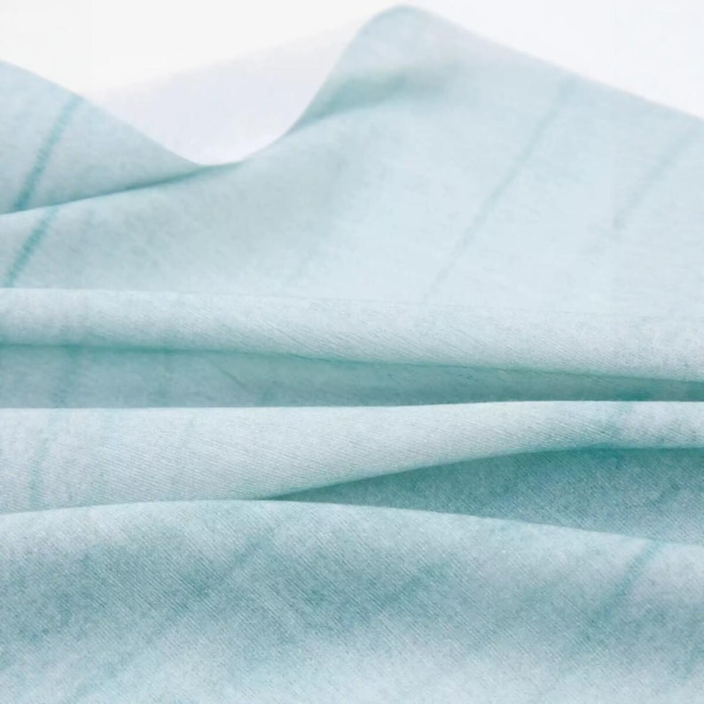 K&ouml;nnte beinhalten: Nahaufnahme eines weichen, hellblauen Stoffes mit dezenten, dunkleren blauen Linien. Der Stoff ist drapiert und gefaltet, wodurch ein strukturiertes Aussehen entsteht. Das Material wirkt leicht und eignet sich m&ouml;glicherweise f&uuml;r Kleidung oder Heimdekoration.