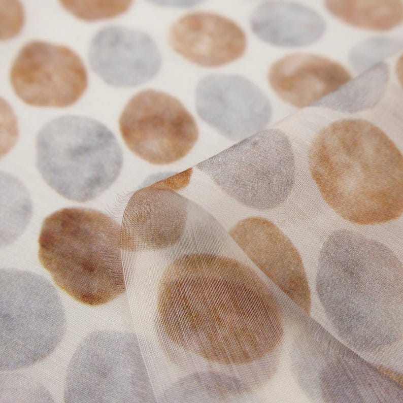 May include: Off-white fabric with a repeating pattern of large, watercolor-style ovals in shades of brown and gray. The fabric is slightly translucent, with a soft, flowing drape. The design is reminiscent of pebbles or organic shapes.