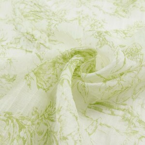 May include: Close-up of a white fabric with a delicate green floral pattern. The fabric is soft and flowing, with a subtle texture. The design features detailed botanical illustrations, creating a fresh and natural aesthetic.