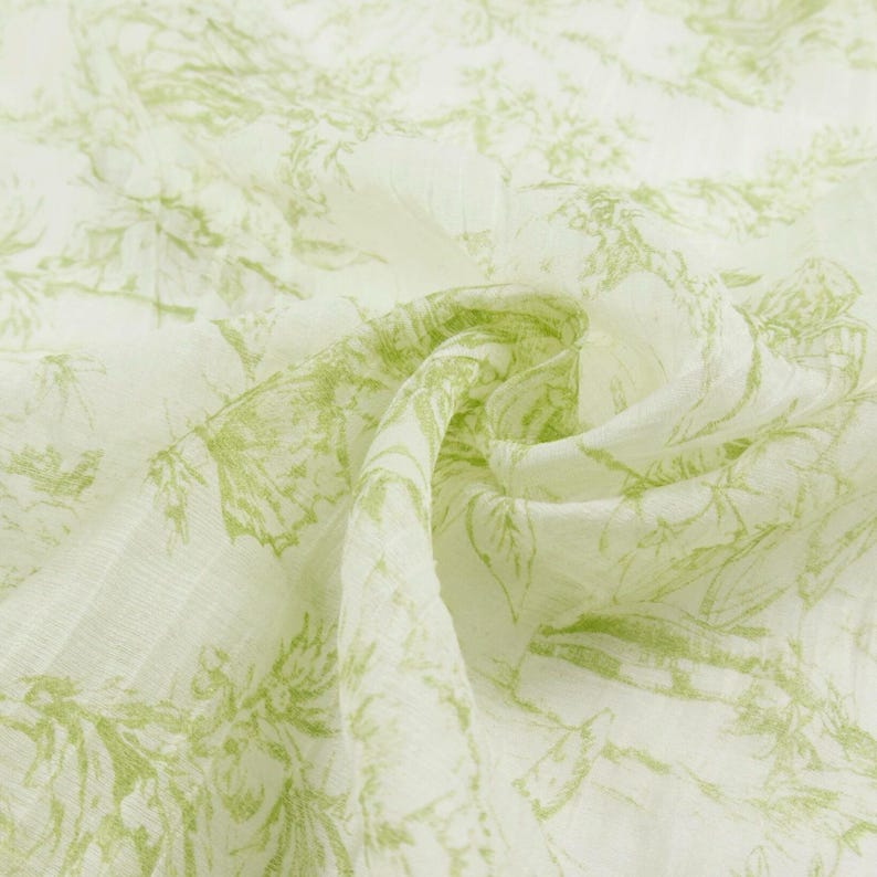 May include: Close-up of a white fabric with a delicate green floral pattern. The fabric is soft and flowing, with a subtle texture. The design features detailed botanical illustrations, creating a fresh and natural aesthetic.