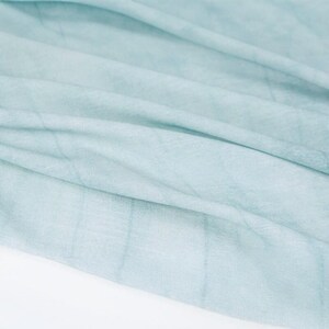 K&ouml;nnte beinhalten: Nahaufnahme eines hellblauen Stoffes mit dezenten gitterartigen Mustern. Der Stoff ist weich und flie&szlig;end, mit sanften Falten und Schatten, die ein Gef&uuml;hl von Tiefe und Textur erzeugen. Das Gesamtbild ist zart und luftig.