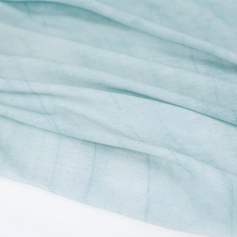 K&ouml;nnte beinhalten: Nahaufnahme eines hellblauen Stoffes mit dezenten gitterartigen Mustern. Der Stoff ist weich und flie&szlig;end, mit sanften Falten und Schatten, die ein Gef&uuml;hl von Tiefe und Textur erzeugen. Das Gesamtbild ist zart und luftig.