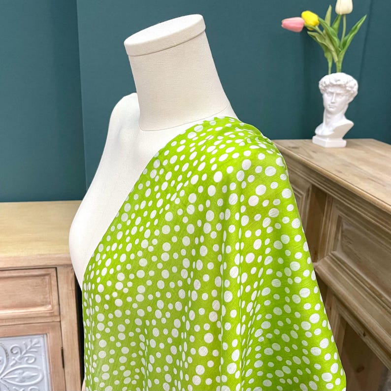 May include: A vibrant lime green fabric featuring a pattern of white polka dots of varying sizes. The textile is draped over a white mannequin torso, showcasing the lightweight texture and the consistent distribution of the circular white print.
