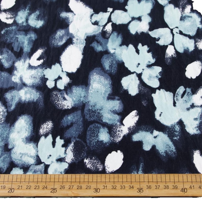 May include: Close-up of a fabric with a floral pattern. The design features light blue and white flowers against a dark navy blue background. A wooden ruler with inch markings is at the bottom.