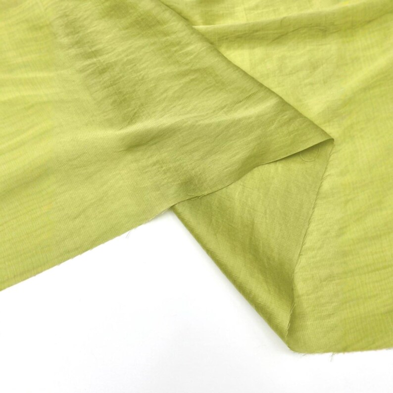 May include: A piece of lime green fabric with a visible woven texture and a slight sheen. The material is draped to show folds and creases, highlighting the light, semi-translucent quality of the textile. The edges appear raw and unfinished.