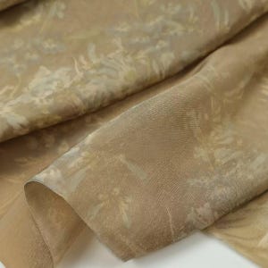 Puede incluir: Una tela ligera y transparente en tonos beige o marr&oacute;n claro, con un sutil estampado floral tono sobre tono. El material est&aacute; drapeado en pliegues suaves, resaltando su cualidad transl&uacute;cida y el delicado dise&ntilde;o bot&aacute;nico repetitivo.