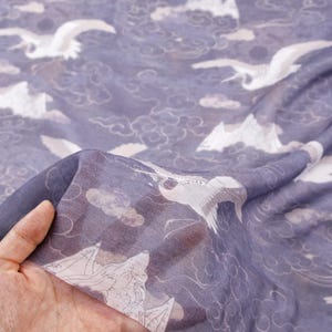 May include: A sheer fabric with a repeating pattern of white cranes, clouds, and mountains on a muted blue background. The fabric is held up, showing its delicate, translucent quality. The design evokes a sense of serenity.