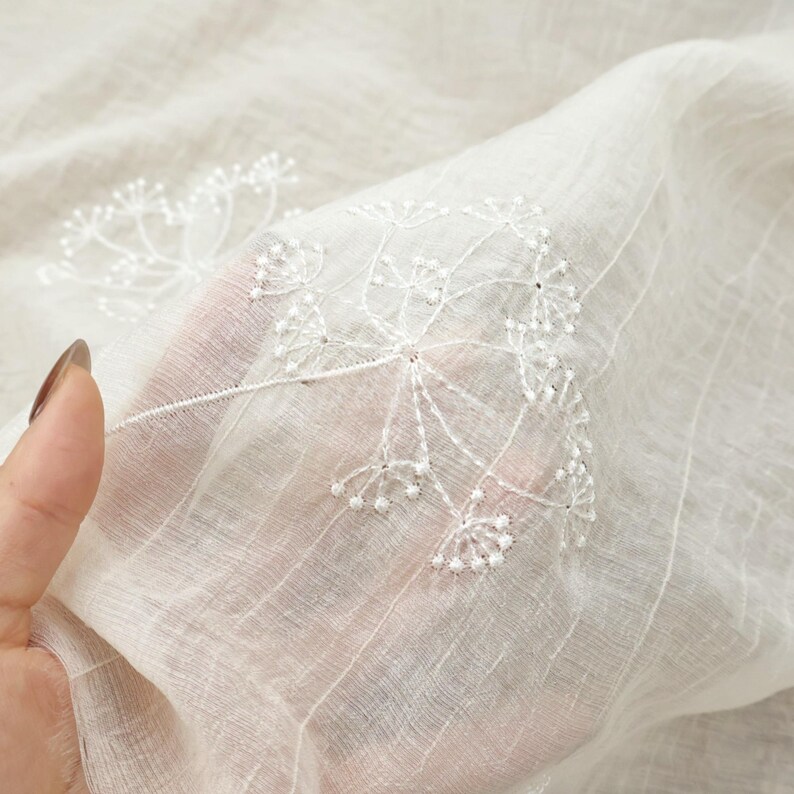 May include: Close-up of sheer white fabric with delicate, embroidered floral patterns. The embroidery features detailed white threadwork, creating a textured, airy design. The fabric has a subtle striped texture, adding depth to the overall aesthetic.