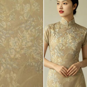 May include: A beige, short-sleeved dress with a mandarin collar and floral pattern. The dress features a wrap-style closure and is adorned with a delicate floral print in shades of white and blue. The fabric has a subtle sheen.