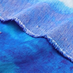 May include: Close-up of a blue fabric with a frayed edge. The fabric has a watercolor effect with shades of blue and hints of purple. The texture of the fabric is visible, with a slightly rough appearance.
