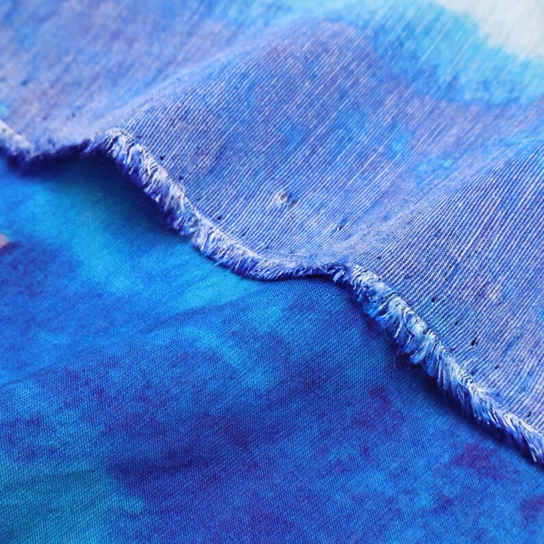 May include: Close-up of a blue fabric with a frayed edge. The fabric has a watercolor effect with shades of blue and hints of purple. The texture of the fabric is visible, with a slightly rough appearance.
