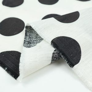 May include: White fabric with large black polka dots. The fabric is folded, revealing a textured weave and frayed edges. The polka dots are evenly spaced and create a bold, graphic pattern. Ideal for home decor or fashion projects.