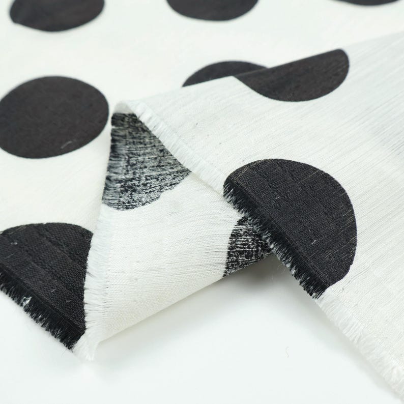 May include: White fabric with large black polka dots. The fabric is folded, revealing a textured weave and frayed edges. The polka dots are evenly spaced and create a bold, graphic pattern. Ideal for home decor or fashion projects.