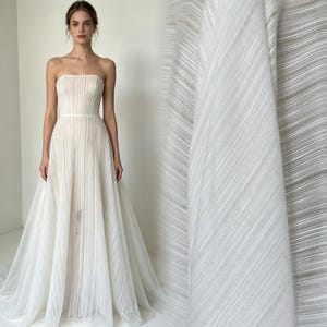 Puede incluir: Un vestido largo sin tirantes con corpiño ajustado y falda fluida, confeccionado en tela semitransparente de color blanco roto con una fina textura plisada vertical. El diseño presenta una cintura definida y una silueta en forma de A.