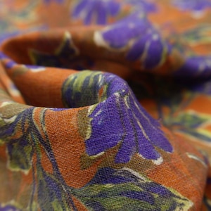 May include: Close-up of a fabric with a floral pattern. The fabric features large purple flowers with green leaves and white accents against a burnt orange background. The fabric is folded, showing texture and depth.