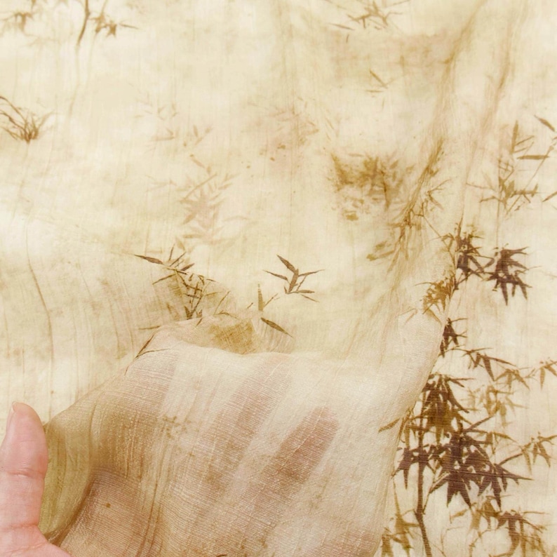 May include: A sheer, beige fabric with a bamboo leaf pattern in shades of brown. The fabric is held up, showing its transparency. The bamboo design is repeated across the surface, creating a natural, organic aesthetic.