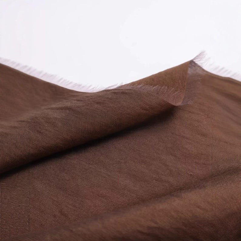 K&ouml;nnte beinhalten: Ein St&uuml;ck dunkelbrauner Stoff mit einer sichtbaren Webstruktur und einer ausgefransten Kante. Das Material ist so drapiert, dass Falten und Schatten auf der Oberfl&auml;che entstehen, was die tiefbraune Farbe und die feine Webart hervorhebt.