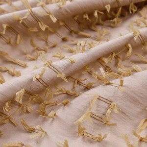 May include: A light beige fabric featuring a textured surface with numerous small, frayed, golden-yellow thread accents arranged in a grid-like pattern. The material is folded, creating soft, flowing drapes across the entire surface area.