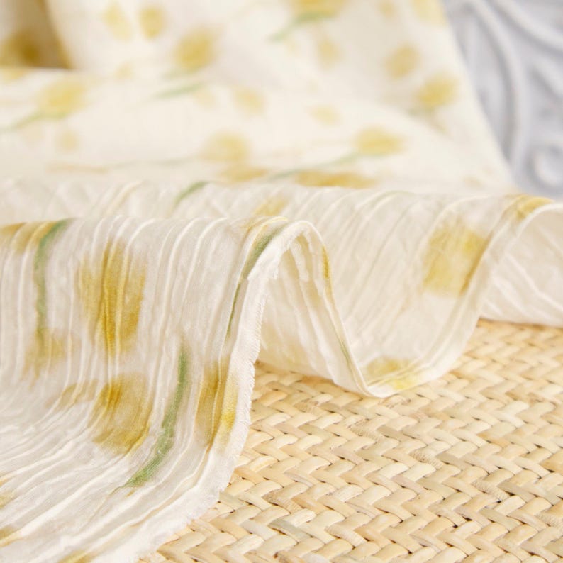 May include: A piece of off-white fabric with a crinkled, textured surface featuring a repeating pattern of soft yellow floral shapes and faint green stems. The material is draped in loose folds, showcasing the lightweight and pleated quality.