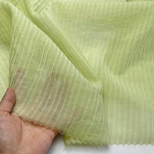 May include: A close-up of a light green fabric with vertical ribbed texture. The fabric is translucent and appears soft, with a slight sheen. The material is being held, showcasing its drape and texture.