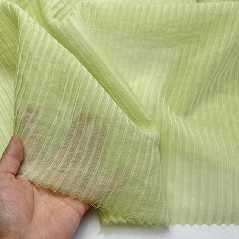 May include: A close-up of a light green fabric with vertical ribbed texture. The fabric is translucent and appears soft, with a slight sheen. The material is being held, showcasing its drape and texture.