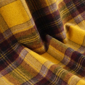 May include: A fabric featuring a plaid pattern in shades of mustard yellow, deep burgundy, and dark olive green. The material has a soft, slightly fuzzy texture with visible folds and drapes that create depth and shadows across the surface.