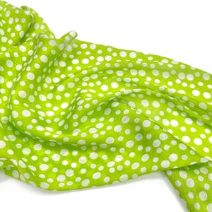May include: A lightweight lime green scarf featuring a pattern of white polka dots of varying sizes. The fabric is draped in soft folds, showcasing the bright green background and the scattered white circular print across the entire surface.