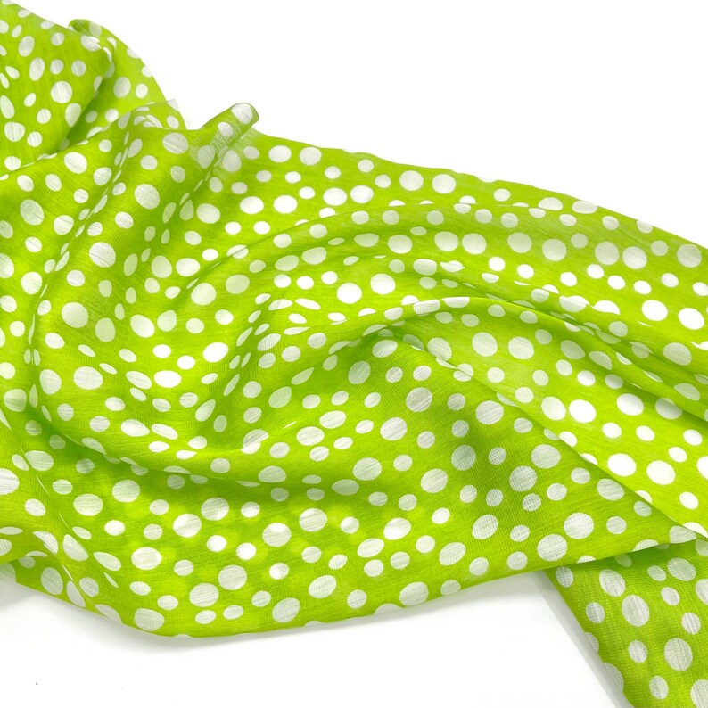 May include: A lightweight lime green scarf featuring a pattern of white polka dots of varying sizes. The fabric is draped in soft folds, showcasing the bright green background and the scattered white circular print across the entire surface.