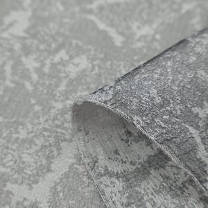 May include: Close-up of a gray fabric with a textured, abstract pattern. The fabric is folded over, revealing the edge and the reverse side. The pattern features light and dark gray tones, creating a mottled effect.