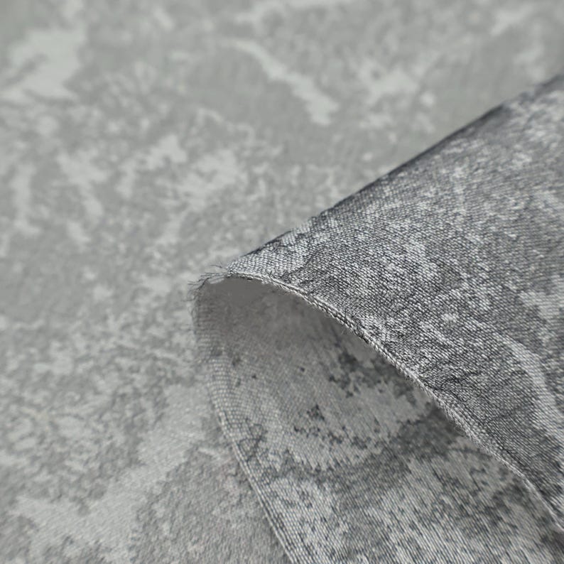 May include: Close-up of a gray fabric with a textured, abstract pattern. The fabric is folded over, revealing the edge and the reverse side. The pattern features light and dark gray tones, creating a mottled effect.