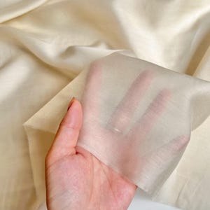 以下が含まれることがあります： 光にかざされた、薄手のクリーム色の生地のクローズアップ。生地は薄く半透明で、わずかにテクスチャ加工された表面です。背景は同じ生地の大きなもので、柔らかい折り目と影があります。