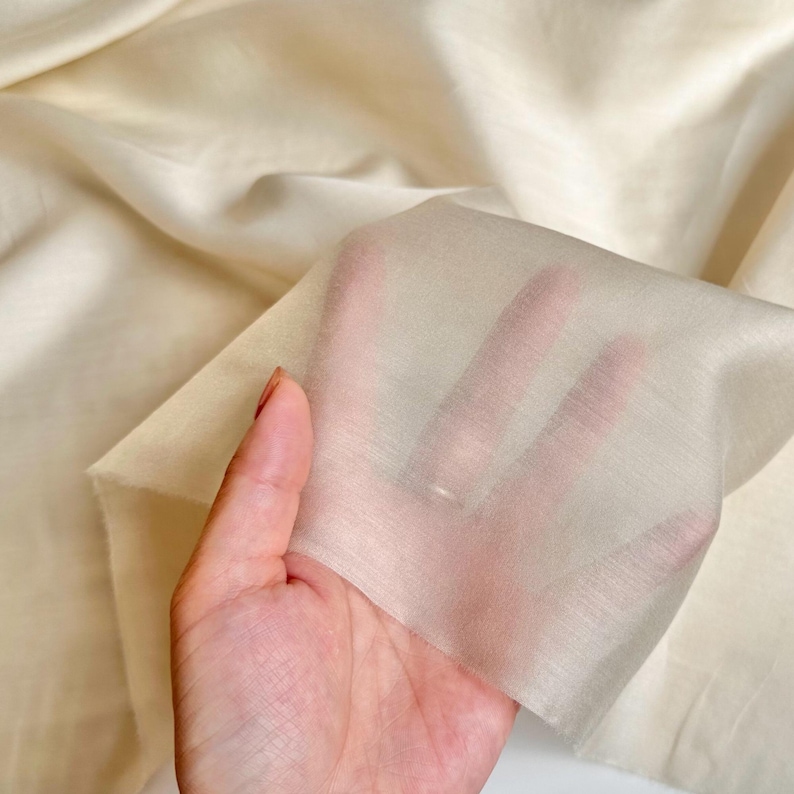 以下が含まれることがあります： 光にかざされた、薄手のクリーム色の生地のクローズアップ。生地は薄く半透明で、わずかにテクスチャ加工された表面です。背景は同じ生地の大きなもので、柔らかい折り目と影があります。