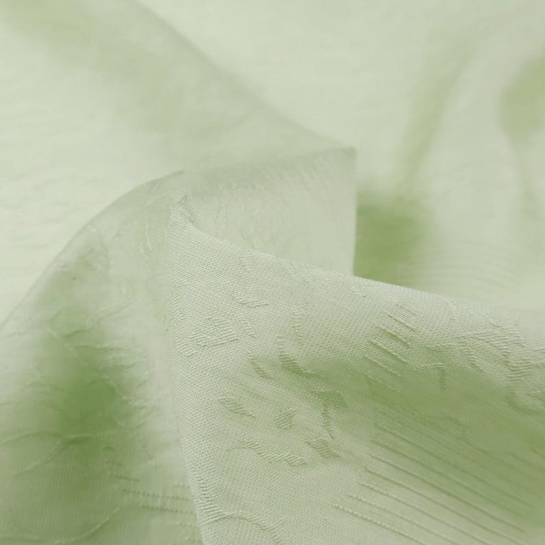 May include: Close-up of a light green fabric with a subtle textured pattern. The fabric appears to be soft and flowing, with gentle folds and highlights. The overall impression is of a delicate, high-quality textile.