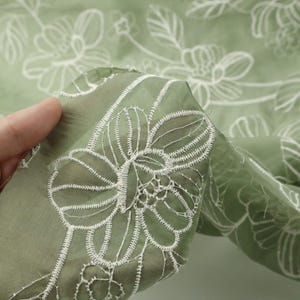 May include: A sheer sage green fabric featuring an intricate white embroidered floral pattern. The delicate textile displays a repeating design of outlined blossoms and leaves, with the embroidery thread creating a raised texture on the surface.
