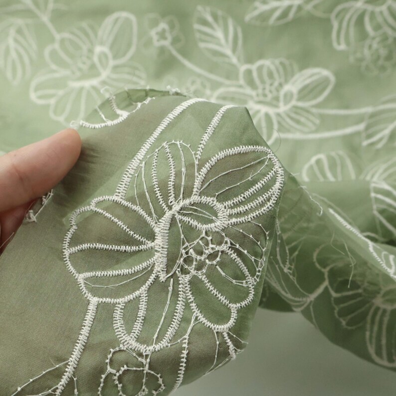 May include: A sheer sage green fabric featuring an intricate white embroidered floral pattern. The delicate textile displays a repeating design of outlined blossoms and leaves, with the embroidery thread creating a raised texture on the surface.
