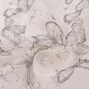 Op de afbeelding: Cr&egrave;me-witte stof met een patroon van grijze vlinderillustraties en verspreide zilveren glitter. De stof is gedrapeerd en toont plooien en textuur. De vlinders zijn getekend met gedetailleerde vleugels.