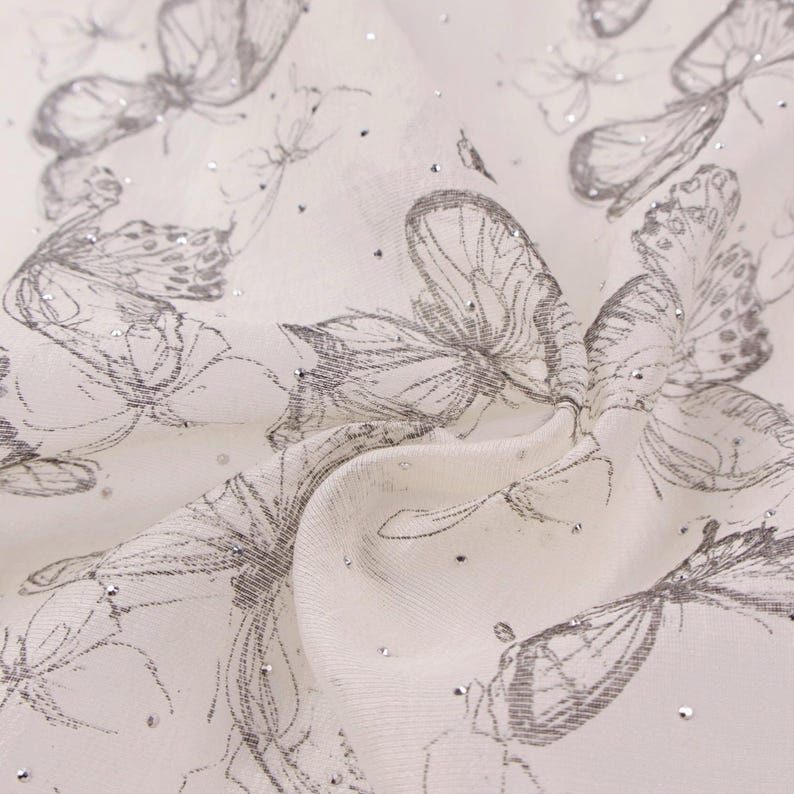 Op de afbeelding: Cr&egrave;me-witte stof met een patroon van grijze vlinderillustraties en verspreide zilveren glitter. De stof is gedrapeerd en toont plooien en textuur. De vlinders zijn getekend met gedetailleerde vleugels.