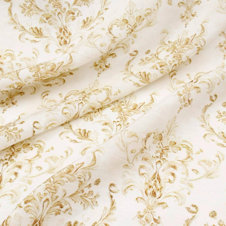 May include: Close-up of a cream-colored fabric with a repeating gold damask pattern. The fabric is folded, creating shadows and highlighting the texture of the material. The design features ornate floral and scroll motifs.