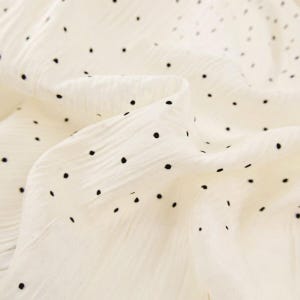 May include: Close-up of a cream-colored fabric with a textured surface and scattered black polka dots. The fabric is folded, creating shadows and highlighting the texture. The overall effect is soft and delicate.