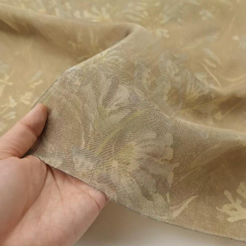 May include: Tan fabric with a floral pattern. The fabric is held up, showing its texture and the details of the design. The floral pattern is in shades of white and green.