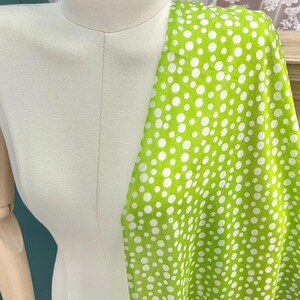 May include: A bright lime green fabric featuring a pattern of white polka dots of varying sizes. The fabric is draped over a neutral-colored mannequin torso, showcasing the contrast between the vibrant green background and the white circular spots.