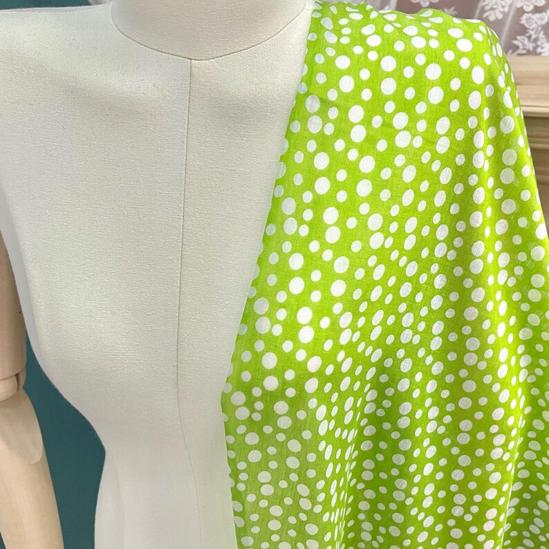 May include: A bright lime green fabric featuring a pattern of white polka dots of varying sizes. The fabric is draped over a neutral-colored mannequin torso, showcasing the contrast between the vibrant green background and the white circular spots.