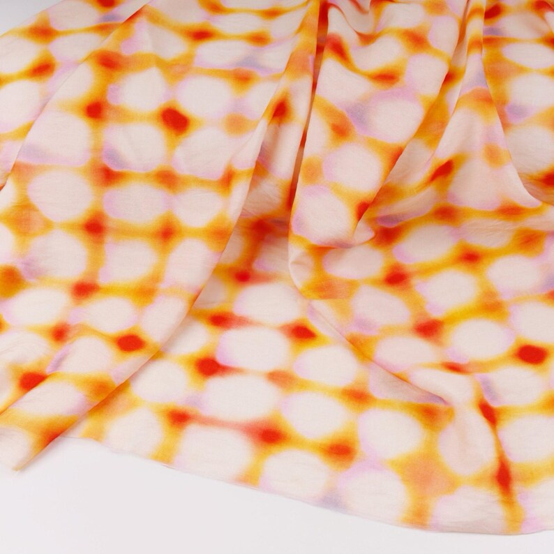 May include: A folded fabric with a repeating pattern of white, orange, and red circles. The fabric has a soft, flowing appearance, with the colors blending together. The background is white.