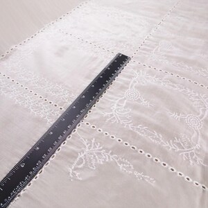 Op de afbeelding: Wit geborduurde stof met bloemen- en geometrische patronen. Een zwarte liniaal, met metingen in centimeters, ligt diagonaal over de stof. De stof heeft delicate uitsparingen en decoratieve stiksels, ideaal voor knutselen of woondecoratie.