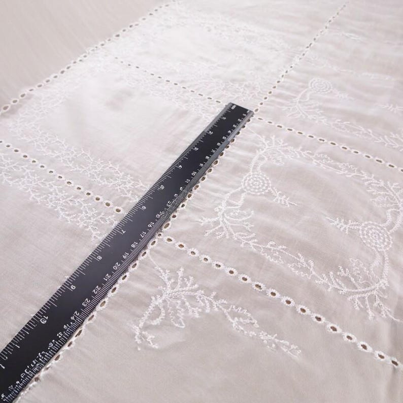 Op de afbeelding: Wit geborduurde stof met bloemen- en geometrische patronen. Een zwarte liniaal, met metingen in centimeters, ligt diagonaal over de stof. De stof heeft delicate uitsparingen en decoratieve stiksels, ideaal voor knutselen of woondecoratie.
