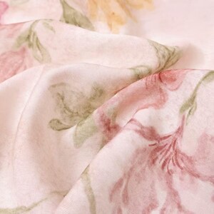 May include: Close-up of a fabric with a floral pattern. The fabric is a light pink color with large, stylized flowers in shades of pink, red, yellow, and green. The fabric is folded, creating shadows and depth, and the overall effect is soft and delicate.