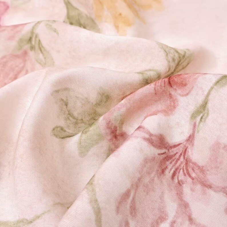 May include: Close-up of a fabric with a floral pattern. The fabric is a light pink color with large, stylized flowers in shades of pink, red, yellow, and green. The fabric is folded, creating shadows and depth, and the overall effect is soft and delicate.