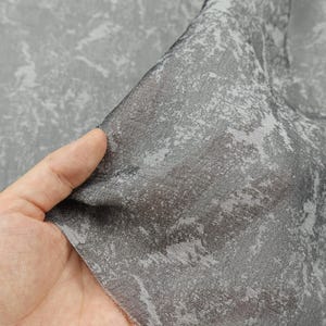 May include: Close-up of a gray fabric with a textured, abstract pattern. The fabric is held up, revealing its semi-sheer quality. The pattern features lighter gray and white accents against a darker gray background, creating a mottled effect. The fabric appears soft and flowing.